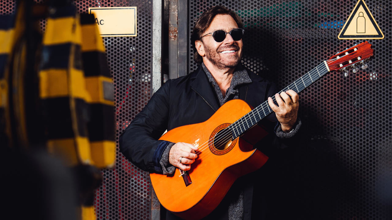 Al Di Meola playing an acoustic guitar backstage at a concert. The background is industrial with metal walls and rough elements.